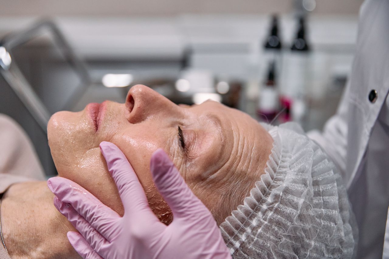 Beautician applies facial treatment on elderly woman with pink gloves in a clinic setting.